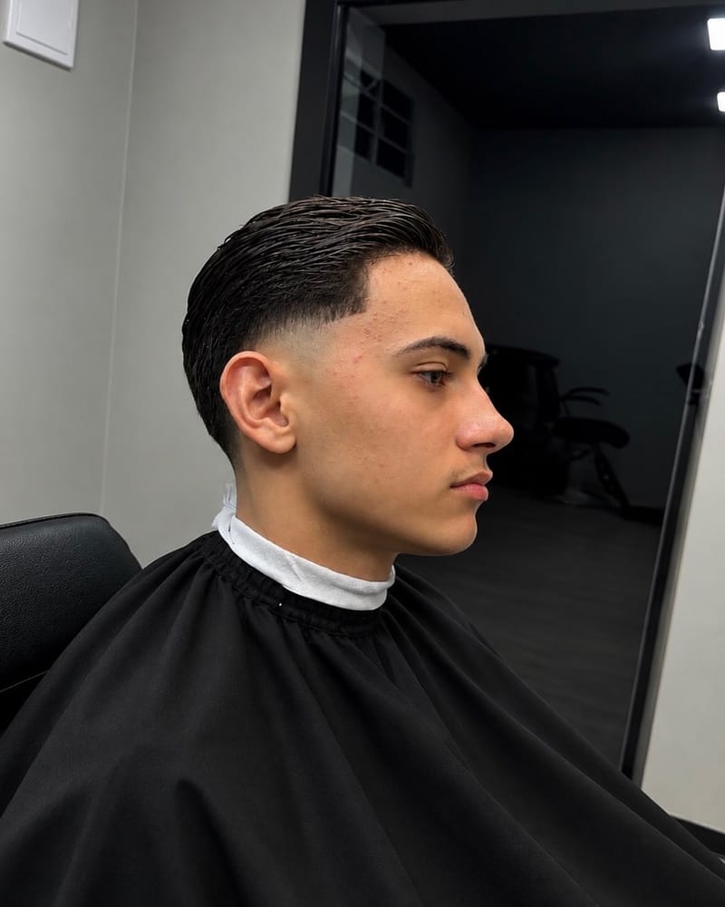 Cliente de perfil com cabelo penteado para trás e nuca com acabamento baixo, de capa preta, na Barbearia União.
