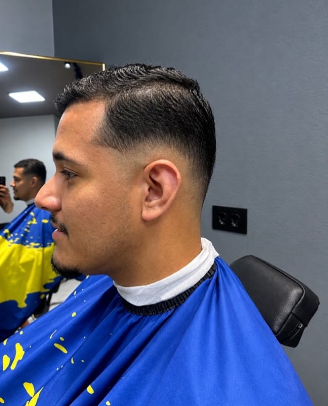Cliente de perfil com cabelo curto penteado para a frente e laterais baixas, usando capa azul e amarela.